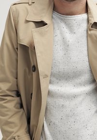 Un trench coat beige con design doppiopetto e bottoni neri, indossato sopra un maglione grigio maculato. Tessuto misto cotone con texture liscia.