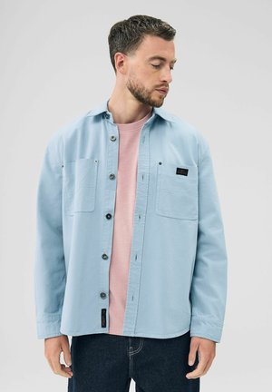 Chaqueta de camisa azul claro de algodón con dos bolsillos en el pecho, cierre de botones y parche de logo negro en el bolsillo izquierdo. Lleva puesta una camiseta rosa debajo.