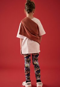 T-shirt bianca, maglione arancione appoggiato sulle spalle, leggings a fantasia colorati, zoccoli bianchi, girata di spalle su uno sfondo rosso.