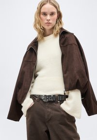 Ξανθιά γυναίκα που φοράει ένα κρεμ αφράτο πουλόβερ, σκούρο καφέ σακάκι ριγμένο στους ώμους, καφέ παντελόνι που ταιριάζει και μαύρη ζώνη με τρουκς, με τα χέρια στις τσέπες.