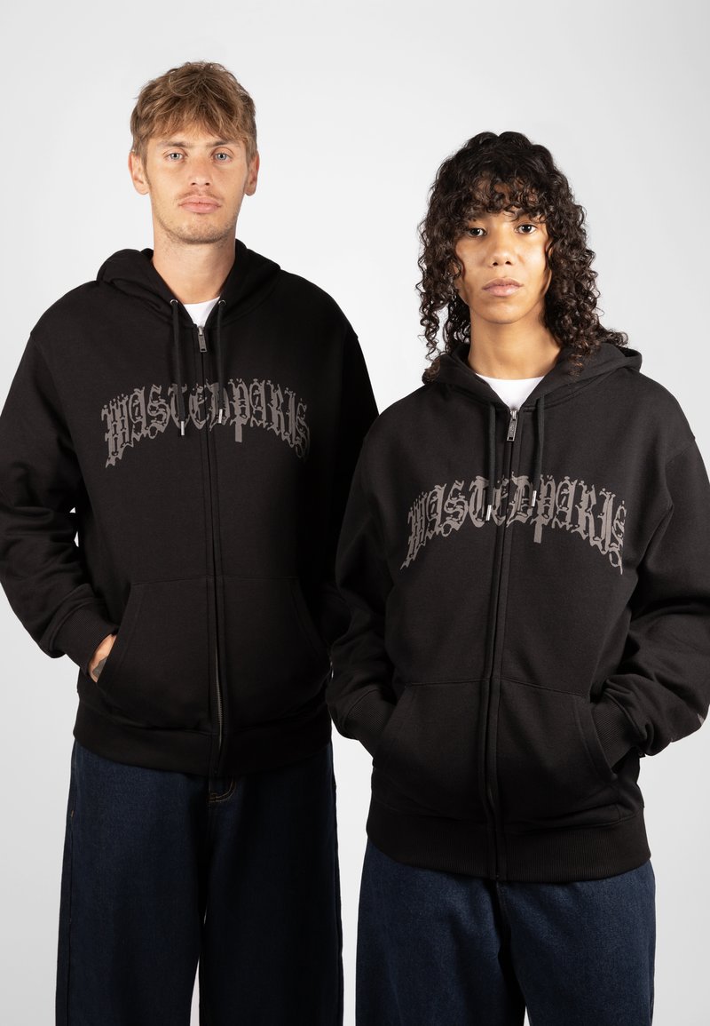 Schwarze Zip-Hoodies mit einem strukturierten, metallisch grauen Logo auf der Vorderseite. Die Modelle tragen lockere Jeanshosen und präsentieren einen lässigen Stil.