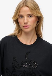 Camiseta de algodón negra con un diseño texturizado y acentos de lentejuelas, cuello redondo y corte relajado. El patrón se asemeja a rasgos animales estilizados.