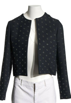 Veste noire courte avec détails en œillets argentés portée sur un mannequin sans tête vêtu d’un pantalon blanc et de colliers superposés.