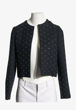 Veste noire courte avec détails en œillets argentés portée sur un mannequin sans tête vêtu d’un pantalon blanc et de colliers superposés.