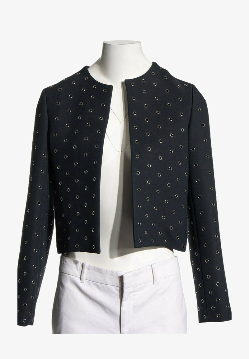 Veste noire courte avec détails en œillets argentés portée sur un mannequin sans tête vêtu d’un pantalon blanc et de colliers superposés.