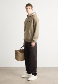 Sudadera caqui con diseño de media cremallera y un bolsillo delantero, combinada con pantalones oscuros y zapatillas blancas, sosteniendo una bolsa de deporte marrón.