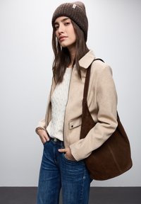 Giacca in misto lana beige con colletto, maglione lavorato a maglia crema, jeans in denim blu e borsa a spalla in suede marrone, indossati con un berretto lavorato a maglia marrone.