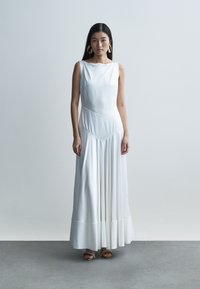 White sleeveless maxi dress with a fitted bodice, pleated skirt, and geometric seams; model wearing large golden hoop earrings.