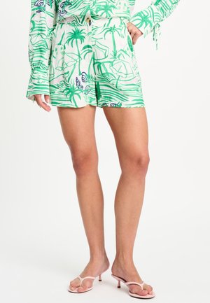 Vrouw draagt groen-witte tropische print korte broek met bijpassend shirt met lange mouwen en roze sandalen met hak.