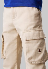 Pantalons cargo beiges en tissu texturé, avec des poches latérales, un petit écusson logo bordeaux et une coupe décontractée.