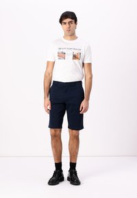 Marineblaue Shorts mit flachem Bund und Taschen, kombiniert mit einem weißen T-Shirt mit grafischem Design und kurzen Ärmeln. Schwarze Schuhe und Socken.