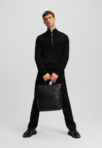 Fekete, cipzáras pulóver bordás gallérral, fekete nadrággal párosítva. A modell egy struktúrált bőrtáska tart, amelyen a "KARL LAGERFELD" felirat van domborítva.