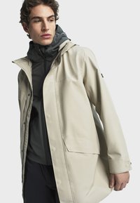 Bež jakna otporna na vodu s kapuljačom, koja ima prednji zip zatvarač, kopču na snapon i velike bočne džepove, preko tamno sive unutarnje jakne.