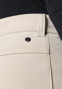 Beige broek heeft een gladde textuur, zichtbare stiksels en een klein zwart metalen knoopaccent op de voorzak.