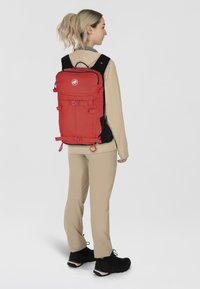 Czerwona plecak wykonany z tkaniny, z wieloma paskami i klamrami. Posiada górny uchwyt i okrągłe logo z przodu. Czarne akcenty.
