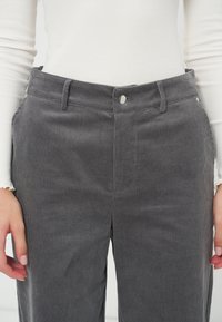 Femme portant un pantalon en velours côtelé gris et un haut blanc à manches longues, mains détendues sur les côtés, taille et hanches visibles.