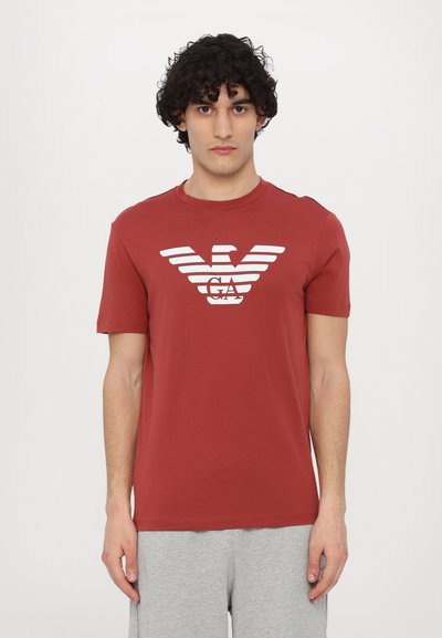 Tricou roșu din bumbac cu un logo alb cu un vultur cu dungi centrat pe piept. Mâneci scurte și guler rotund. Stilizat cu pantaloni scurți gri.