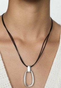 Srebrny owalny wisiorek na czarnym podwójnym sznurku, z matowym wykończeniem i gładką fakturą, noszony na gołej skórze.