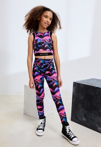 Šarena aktivna odjeća u kompletu koja uključuje kratku majicu i tajice s crnom osnovom te živopisnim vrtložnim uzorcima u ružičastoj i plavoj boji. Crne tenisice s visokim vrhom.