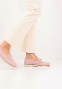 Ρόζ δερμάτινα μοκασίνια με φιόγκο, επίπεδη σόλα και διακριτικές ραφές. Φορεμένα με κρεμ παντελόνια φαρδιάς γραμμής, δείχνοντας ένα casual στυλ.