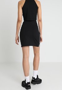 Černý zkrácený halter top, přiléhavá černá mini sukně s bočním logem, doplněná černými sportovními teniskami a bílými ponožkami. Hladká textilní textura.