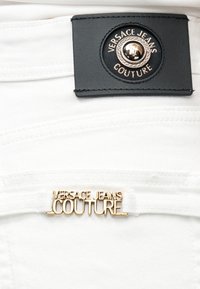 Λευκό ύφασμα denim με χρυσό έμβλημα "Versace Jeans Couture" και μαύρη δερμάτινη ετικέτα με κουμπί λογότυπου σε χρυσό και μαύρο.