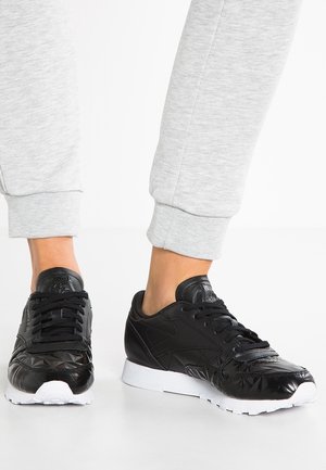 Osoba ubrana w czarne sneakersy Reebok z białymi podeszwami i szare spodnie dresowe stojąca na białej podłodze.