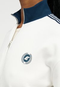 Λευκό μπουφάν με φερμουάρ, με ναυτικό γιακά και ρίγες στον ώμο, διακοσμημένο με ναυτικό λογότυπο που έχει το γράμμα "E" περιβαλλόμενο από φύλλα δάφνης.