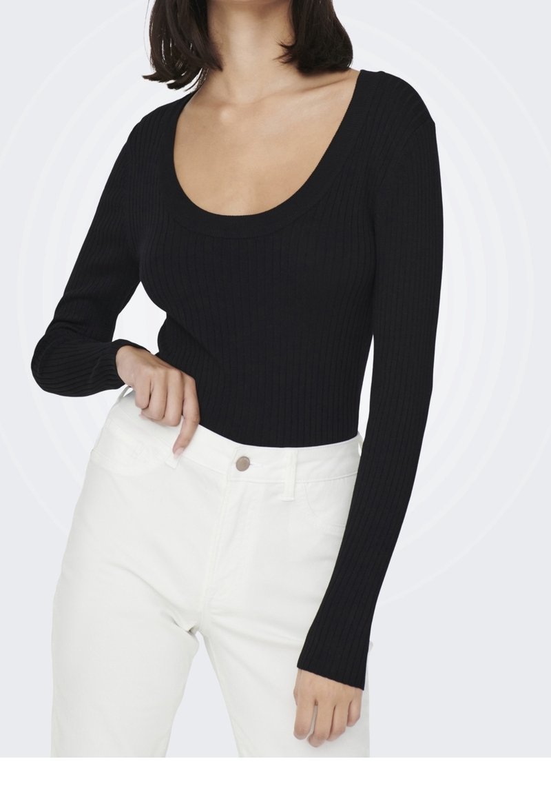 Femme portant un haut noir cintré à manches longues côtelées avec un col rond et un pantalon blanc taille haute, debout devant un fond uni.