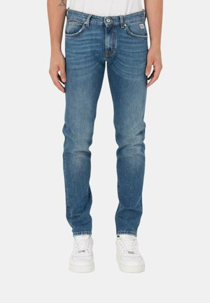 Uomo che indossa jeans blu aderenti e scarpe da ginnastica bianche, in piedi contro uno sfondo chiaro e semplice.