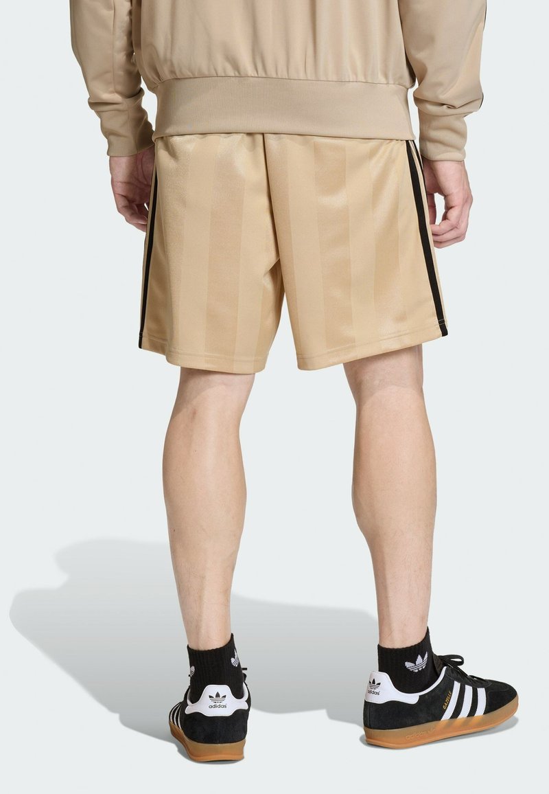 Pantalones cortos deportivos de color beige con rayas verticales, acentos laterales negros, combinados con zapatos negros con logo blanco y suelas de goma. Tela suave.