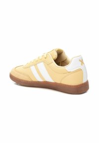 Zapatillas amarillas con rayas blancas, suela de goma texturizada, material superior liso, cuello acolchado y un pequeño acento dorado en el talón.