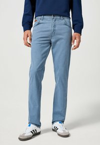 Jeans de pierna recta de color azul claro, hechos de denim, con cierre de botón en la parte delantera y cinco bolsillos. Combinados con zapatillas blancas y una blusa de color azul oscuro.