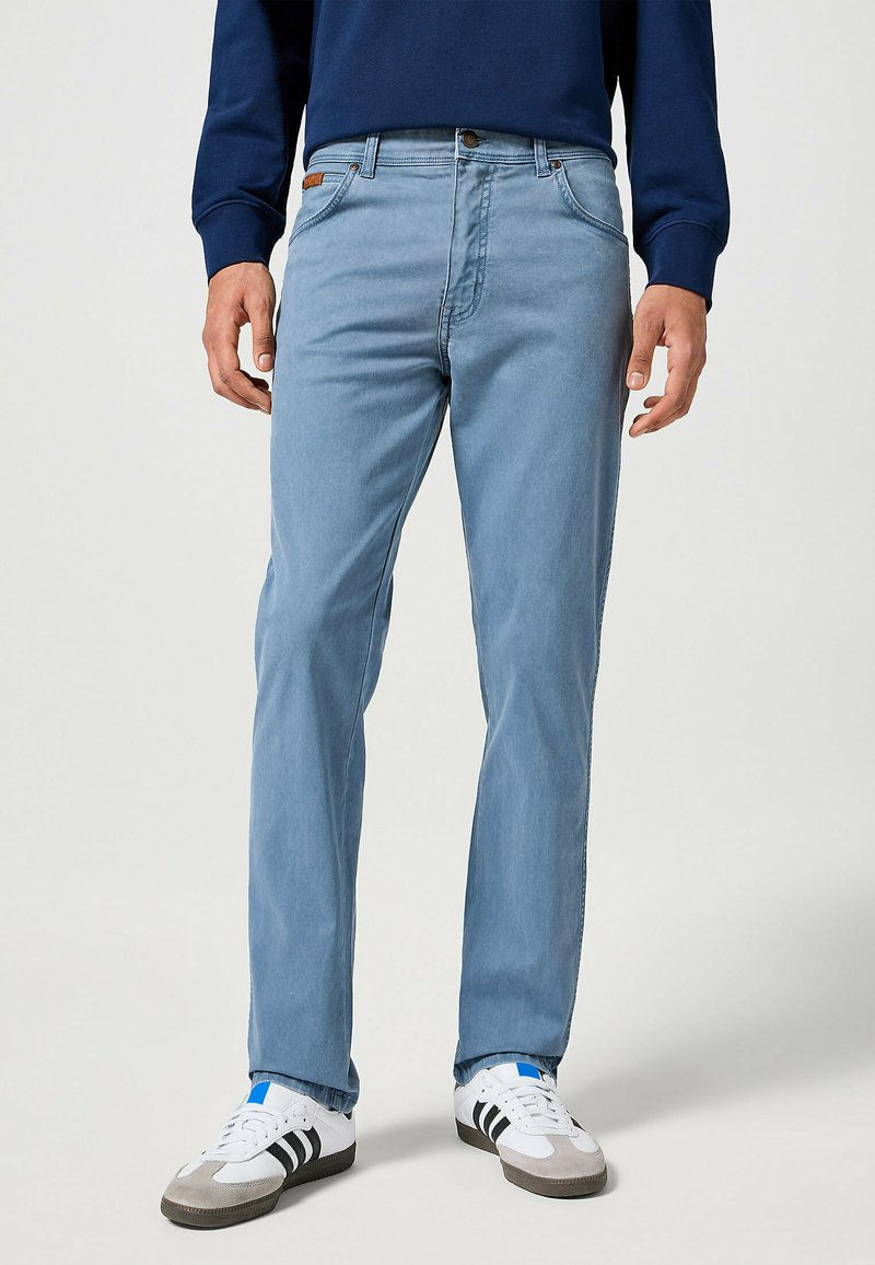 Jeans de pierna recta de color azul claro, hechos de denim, con cierre de botón en la parte delantera y cinco bolsillos. Combinados con zapatillas blancas y una blusa de color azul oscuro.