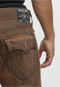 Jeans de mezclilla marrón con un patrón de costura distintivo en el bolsillo trasero y una etiqueta tejida con el nombre de la marca "True Religion."