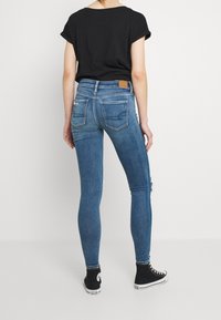Jeans ajustados azules con un acabado desgastado y detalles rasgados, combinados con una blusa negra de mangas enrolladas y zapatillas altas negras.