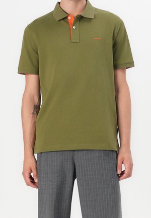 Man wearing olive green polo shirt with orange button placket and small logo, paired with gray pinstriped trousers.