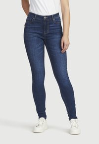 Mörkblå skinny jeans med hög midja, mjuk textur, orange sömmar och en klassisk design med fem fickor, tillsammans med vita sneakers.