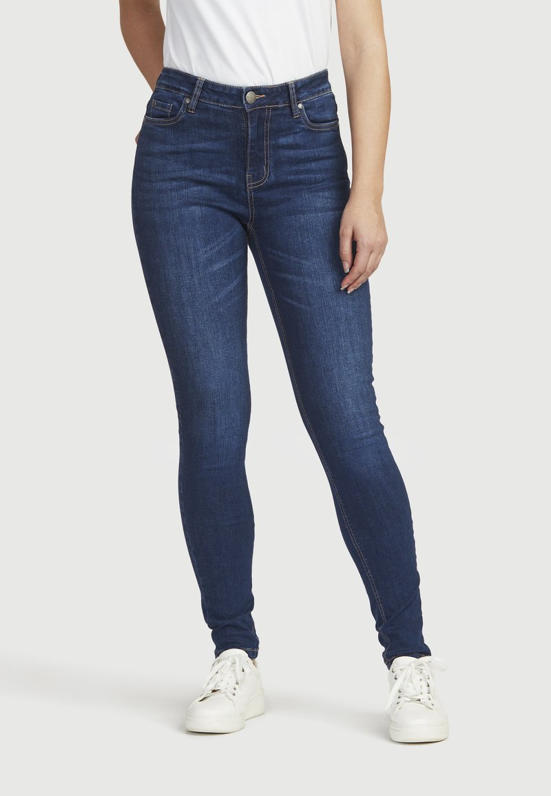 Mörkblå skinny jeans med hög midja, mjuk textur, orange sömmar och en klassisk design med fem fickor, tillsammans med vita sneakers.