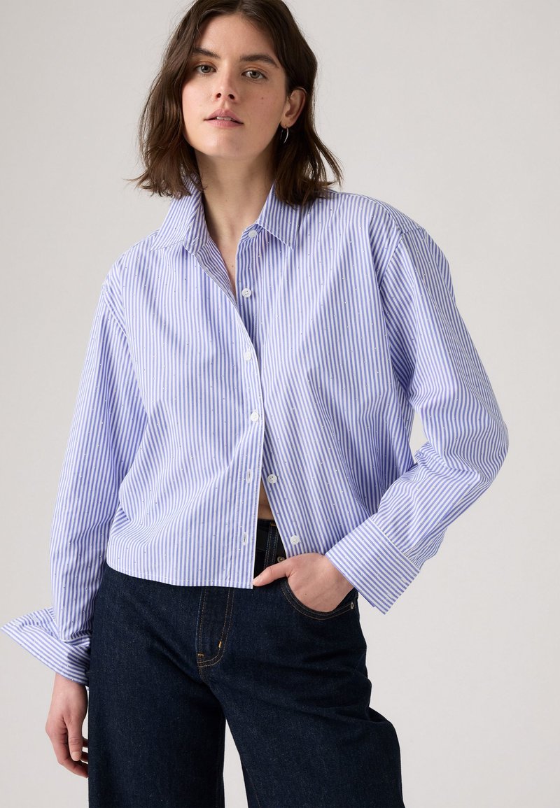 Chemise à rayures bleues et blanches avec un col évasé, des manches longues et une coupe décontractée. Associée à un pantalon en denim foncé.