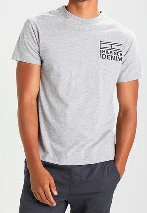 Mężczyzna w jasnoszarej koszulce Tommy Hilfiger Denim i ciemnoszarych spodniach, stoi z jedną ręką w kieszeni, a drugą opuszczoną.