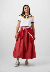 Jonge vrouw staand, gekleed in een wit Duits voetbalshirt, rode rok, rode jas gestrikt rond de taille, witte sokken en zilveren sneakers.