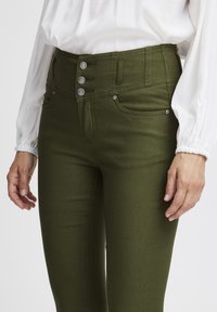 Pantalones de talle alto en verde oliva con tres botones metálicos, bolsillos laterales y una textura suave y elástica. Combinados con una blusa blanca.