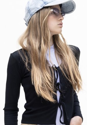 Zwart geribbelde langemouwen cropped top met vetersluiting, gecombineerd met een lichtgrijze denim pet en oversized zonnebril, tegen een witte achtergrond.