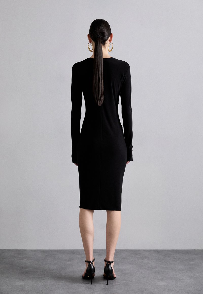 Black, form-fitting long-sleeve dress with a straight silhouette. Smooth texture, knee-length, featuring a seam down the back. High-heeled black sandals.