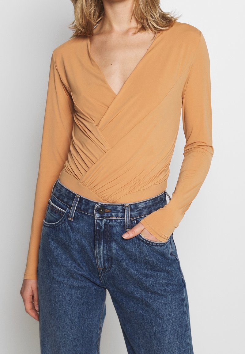 Langärmliges Wickeloberteil aus leichtem, sandfarbenem Stoff mit tiefem V-Ausschnitt, mit gerafften Seiten und kombiniert mit hoch taillierten blauen Jeans.