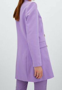 Femme en blazer lavande et pantalon assorti, aux longs cheveux bruns, vue de profil sur un fond clair uni.