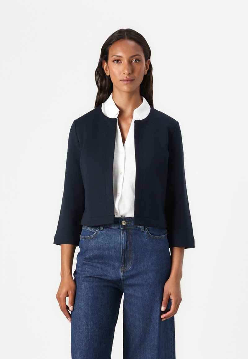 Marineblauwe cropped jas met driekwartmouwen, een getailleerd ontwerp en een ronde halslijn. Gelijk over een wit overhemd met knopen gedragen. Denim broek zichtbaar.