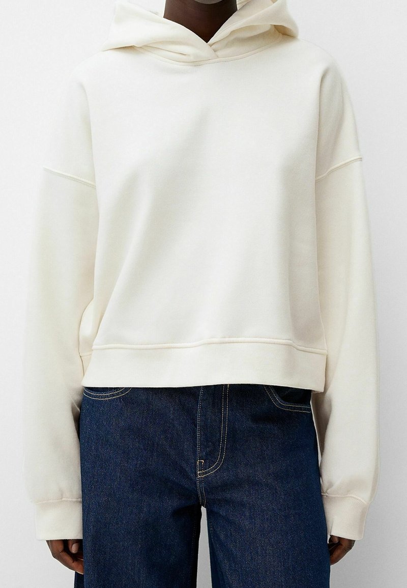 Felpa color crema in tessuto morbido, design corto con un ampio cappuccio, spalle scese e polsini a costine. Indossata con jeans in denim scuri.