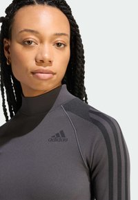 Mladá žena s vrkočmi, oblečená v sivej roláke Adidas s čiernymi pruhmi na ramene a logom Adidas.